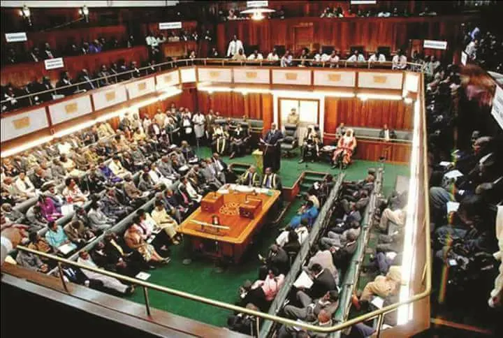 A large group of people in formal attire seated and standing inside a parliamentary chamber with wooden walls, green carpet, and desks, engaged in a legislative session.