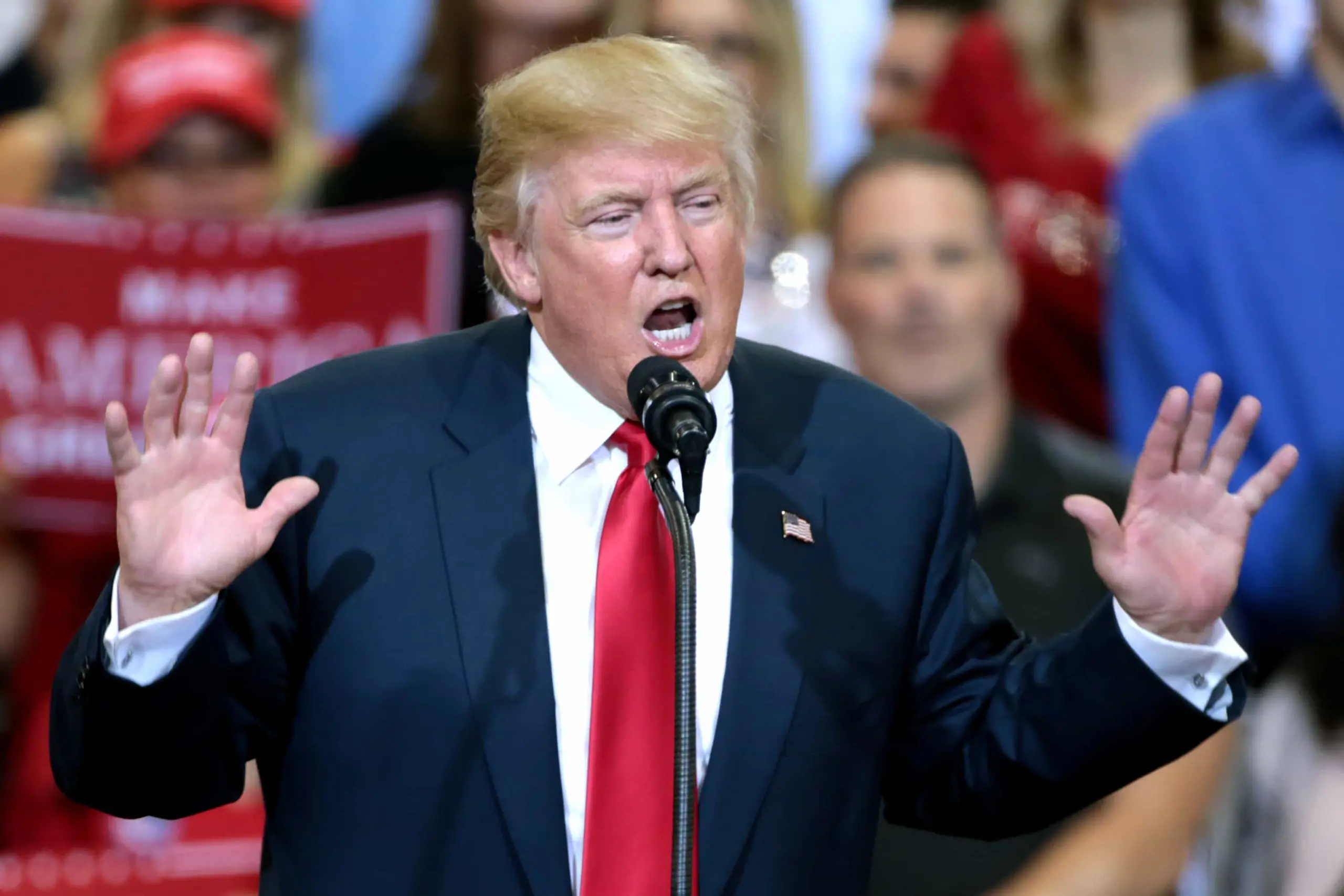 A man in a suit and red tie speaks passionately at a microphone during a rally, addressing how the free press is threatened, with people and campaign signs visible in the blurred background.