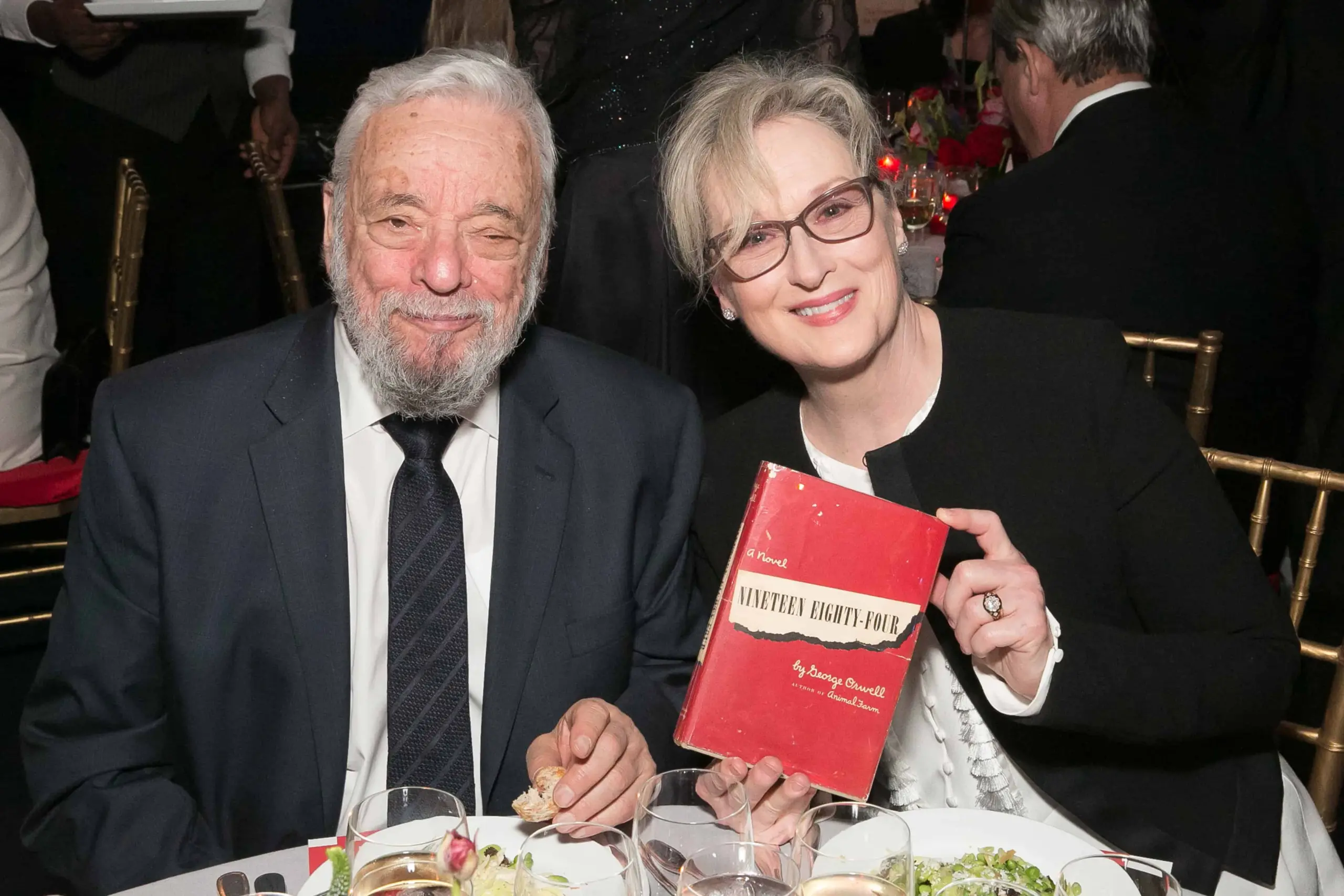 An older man in a suit and a woman in glasses smile at a formal dinner table. The woman holds up a red book titled Nineteen Eighty-Four by George Orwell. Plates with food and drinks are visible on the table.