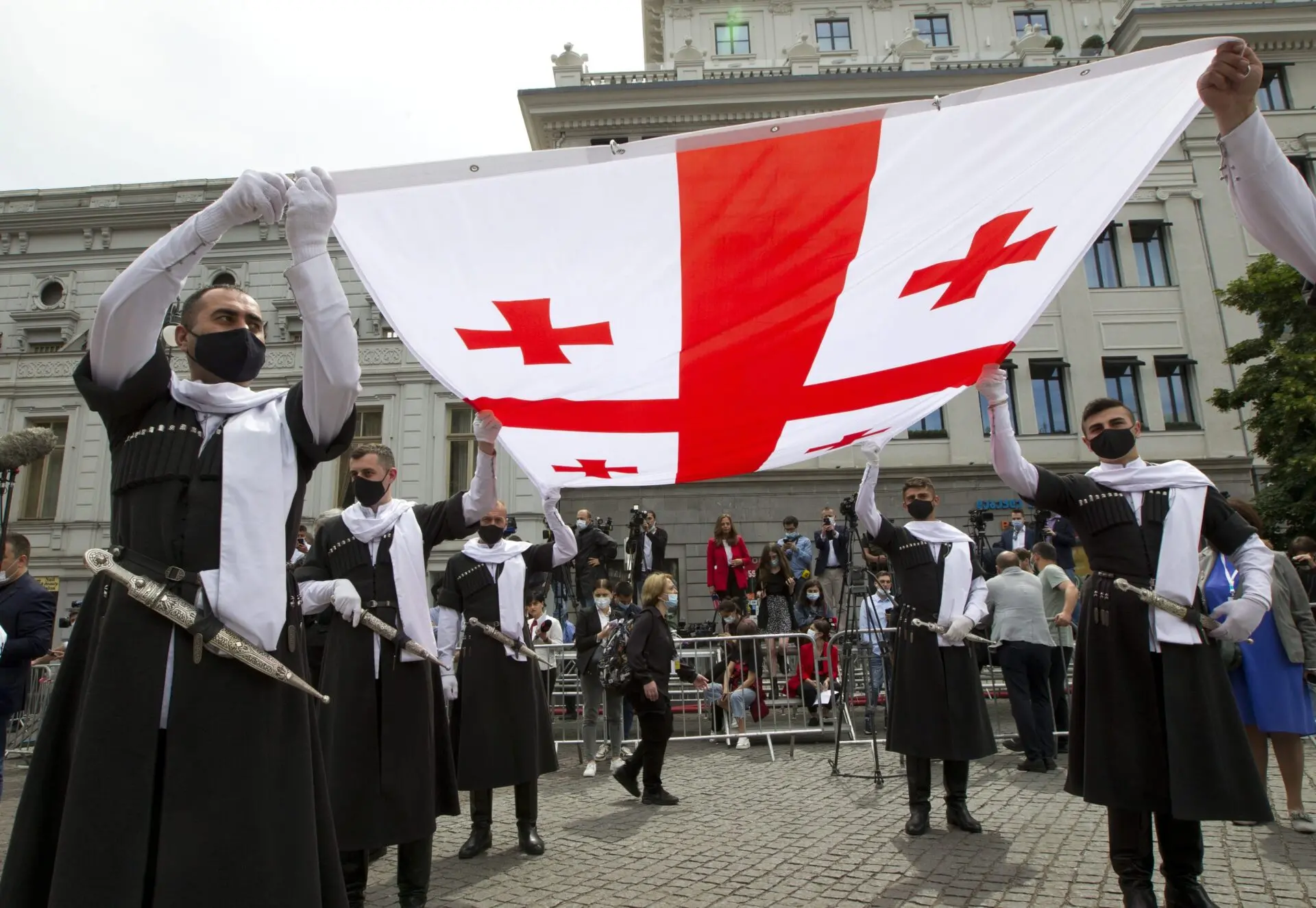 Men in traditional Georgian attire and masks hold up a large Georgian flag in a public square, with a crowd and a historic building in the background.