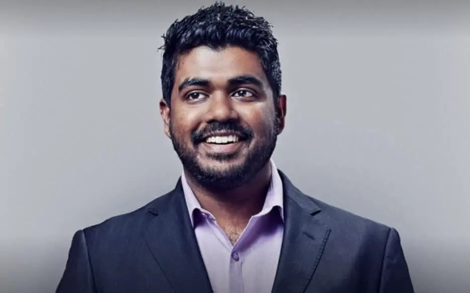 A man with short dark hair and a beard, wearing a dark suit jacket over a light lavender shirt, smiles while looking slightly to the side in front of a plain gray background.