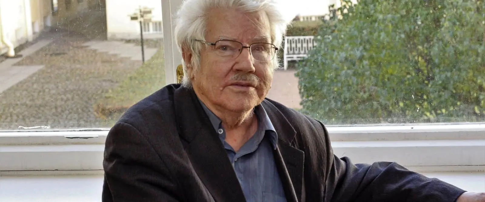 An elderly man with white hair, glasses, and a mustache sits indoors near a window, wearing a dark blazer and blue shirt. Trees and cobblestones are visible outside.