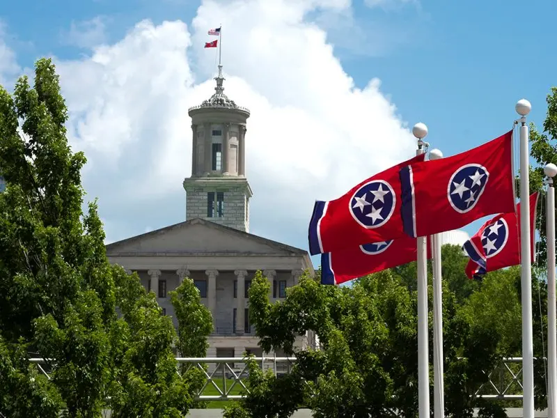 Red Tennessee state flags with three white stars wave in front of the Tennessee State Capitol building, partially obscured by trees, under a blue sky with clouds.