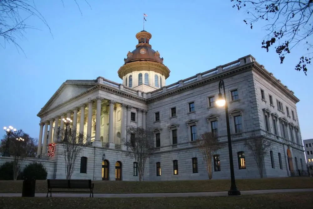 A large historic building with white stone columns and a copper dome, lit up at dusk. Trees and a bench are in the foreground, and the sky is a gradient of light blue.