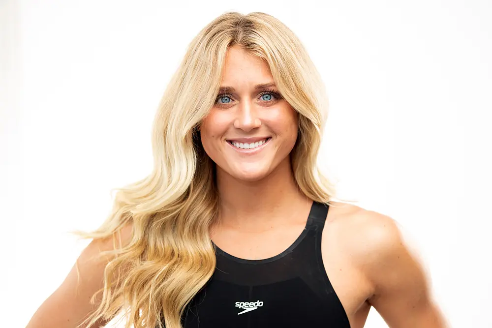 A woman with long blonde hair wearing a black Speedo swimsuit smiles at the camera against a plain white background.