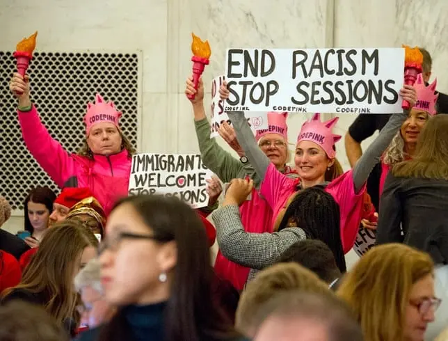 A group of protestors in pink hats and shirts hold signs reading “END RACISM STOP SESSIONS” and “IMMIGRANTS WELCOME,” standing among a seated crowd indoors.