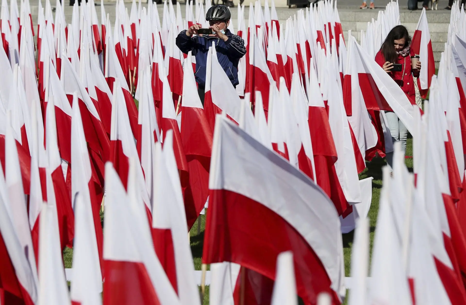 People stand among many red and white flags, with one person taking a photo and another person holding a phone. The flags are arranged closely together outdoors on grass.