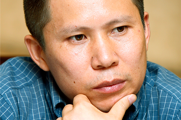A man wearing a blue plaid shirt rests his chin on his hand, looking thoughtful and slightly serious—reminiscent of essayist Xu Zhiyong of China. The background is out of focus.