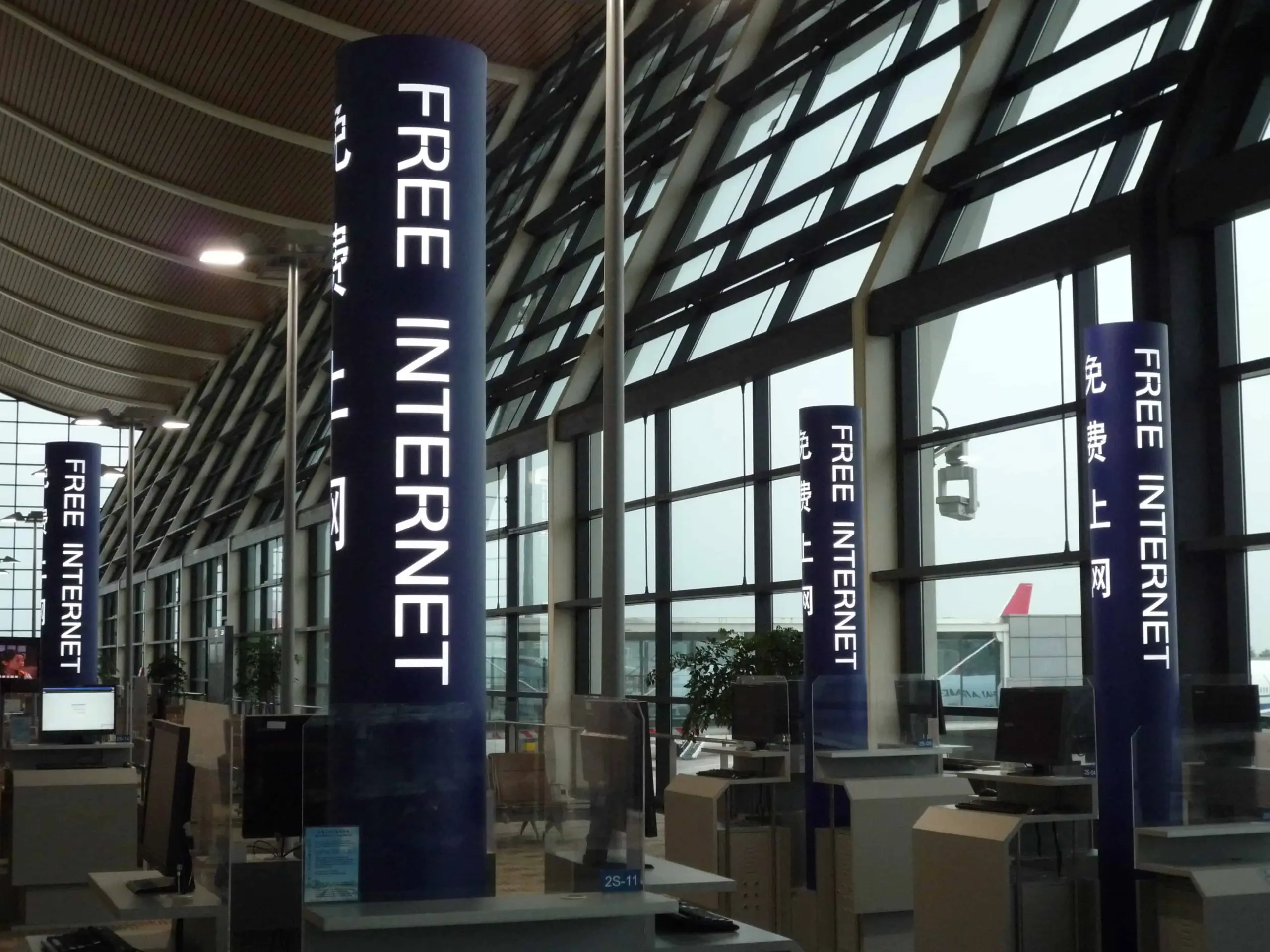 Tall pillars inside an airport terminal display signs reading FREE INTERNET in English and Chinese, with computer stations beneath them and large windows showing part of an airplane outside.