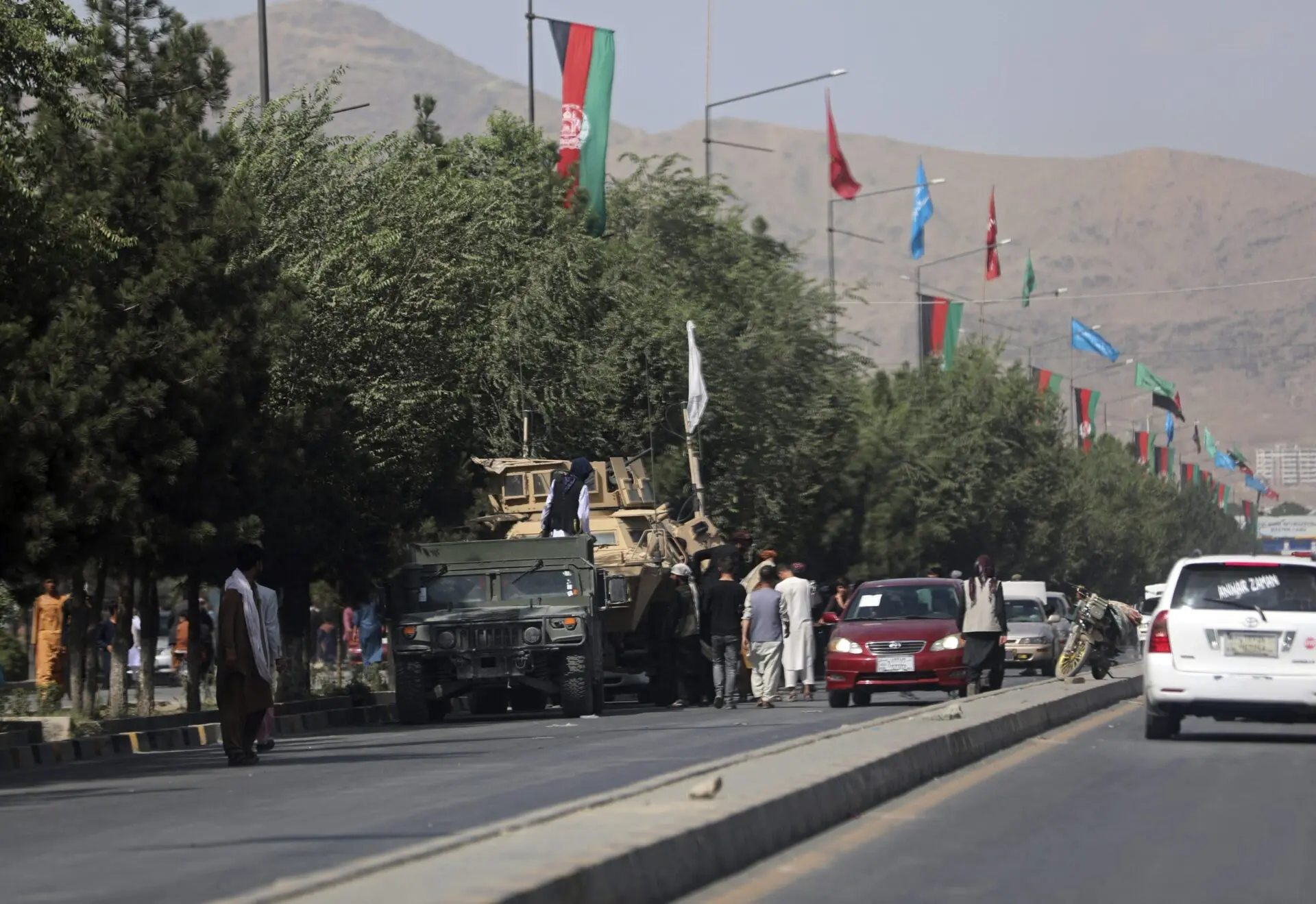 A group of people stands near military vehicles parked on a tree-lined street decorated with Afghanistan flags and banners, with mountains visible in the background. Several cars are also parked along the road.
