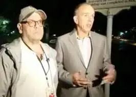 Two men stand outdoors at night; one wears a light jacket, glasses, and a cap, while the other wears a light-colored suit. The scene is dimly lit, and a street sign is visible in the background.