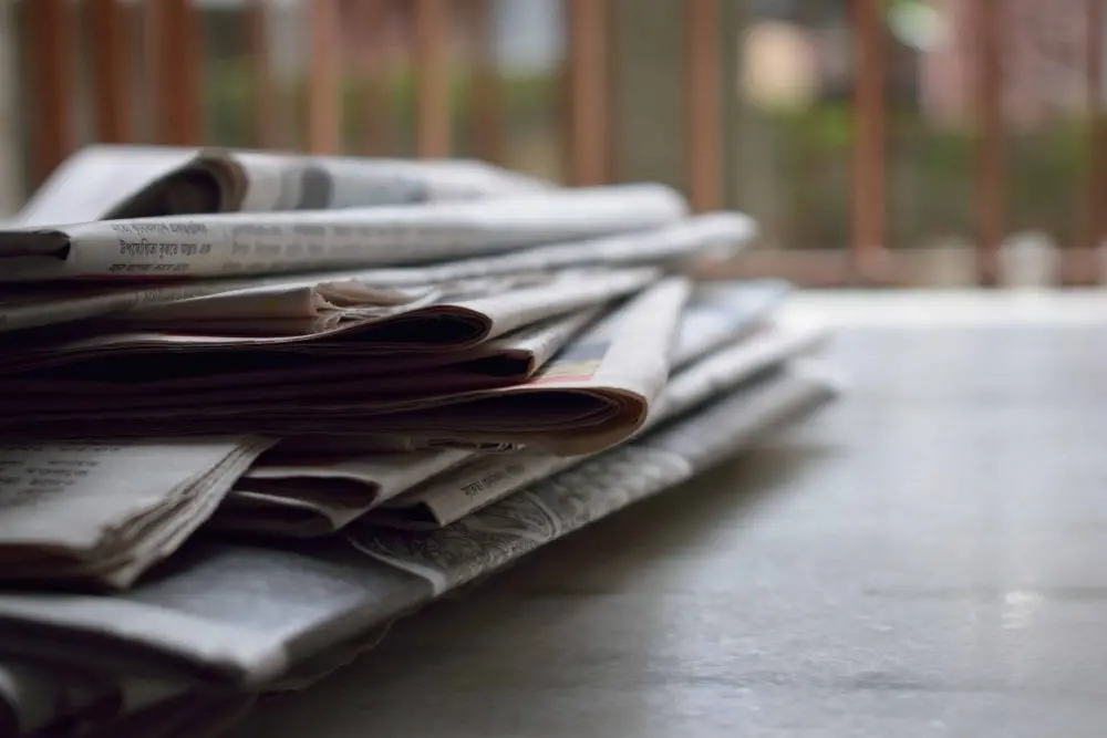 A stack of folded newspapers lies on a smooth surface with a blurred outdoor background, suggesting a quiet, peaceful setting.