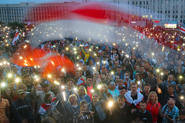 A large crowd gathers at night, waving red-and-white flags and holding up smartphones with lights, creating a sea of illuminated points. The energetic and unified scene appears to protest Belarus leader Lukashenko in front of city buildings.