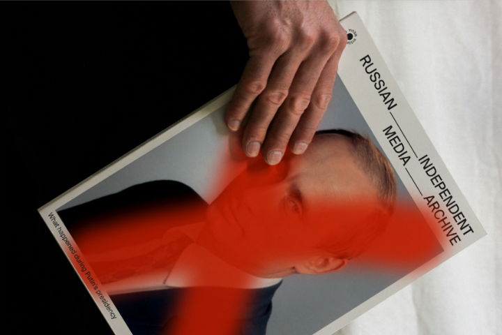 A hand rests on a magazine titled Russian Independent Media Archive featuring a mans portrait, with a bold red X painted across his face.