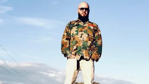 A person with a beard wearing sunglasses, a camouflage jacket, and light-colored pants stands outdoors against a blue sky with light clouds.
