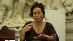 A woman with curly dark hair speaks into a microphone at a panel. She gestures with her hand. Behind her is a white sculpted relief. A water bottle and a glass sit on the table in front of her.