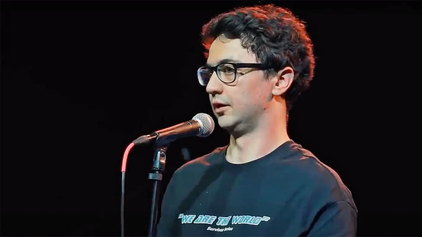 A person with short, dark, curly hair and glasses stands in front of a microphone on stage, wearing a dark T-shirt with white text, against a dark background.
