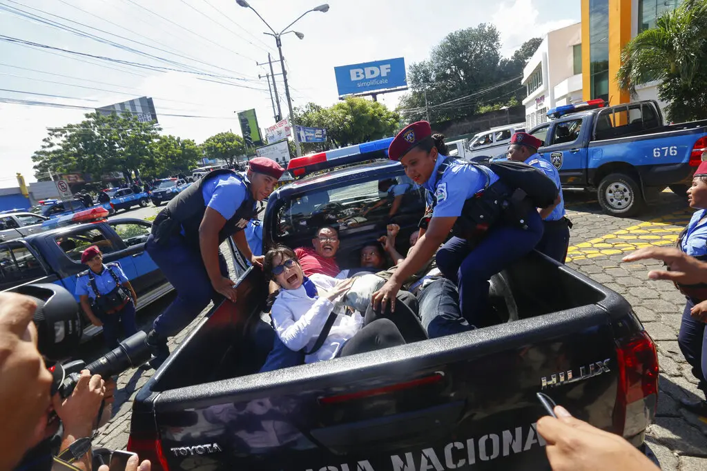 Several police officers detain and forcefully hold down people in the back of a police pickup truck, while others photograph or record the scene on a busy street with more police vehicles in the background.