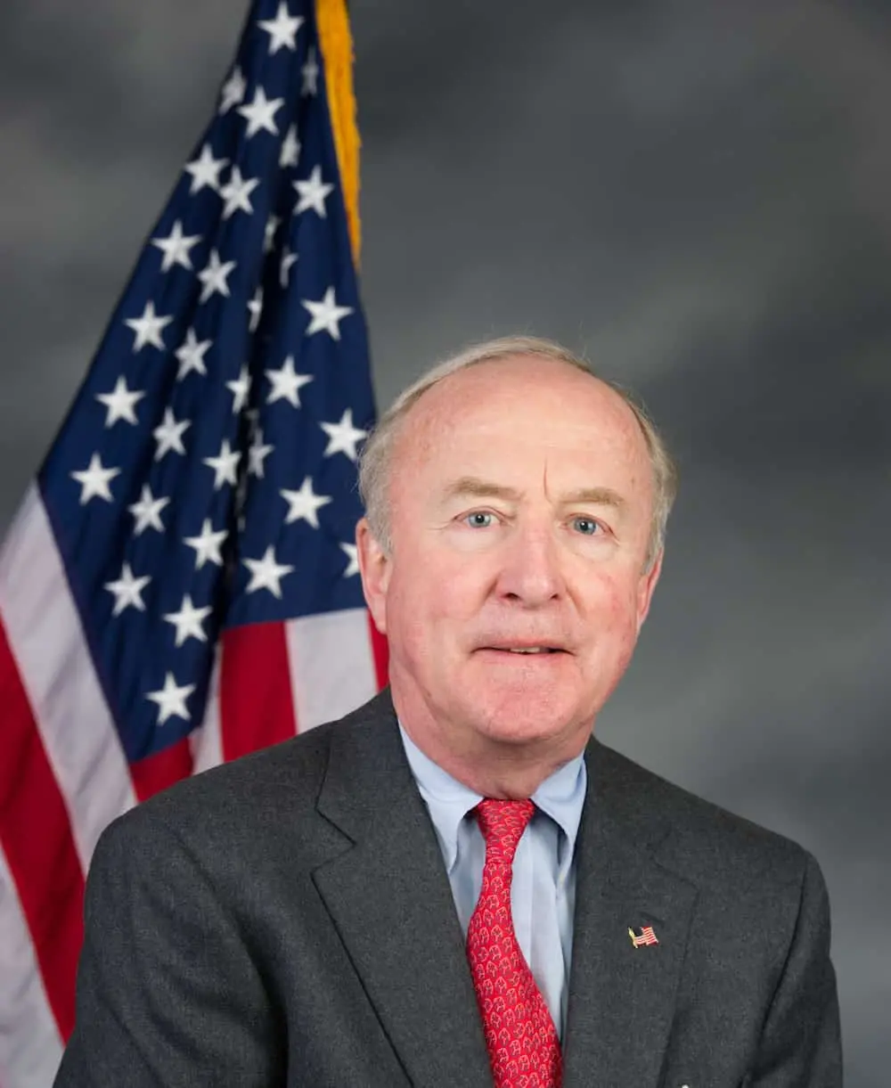 A man in a dark suit and red tie is posing in front of an American flag, with a neutral gray background. He is looking directly at the camera with a slight smile.