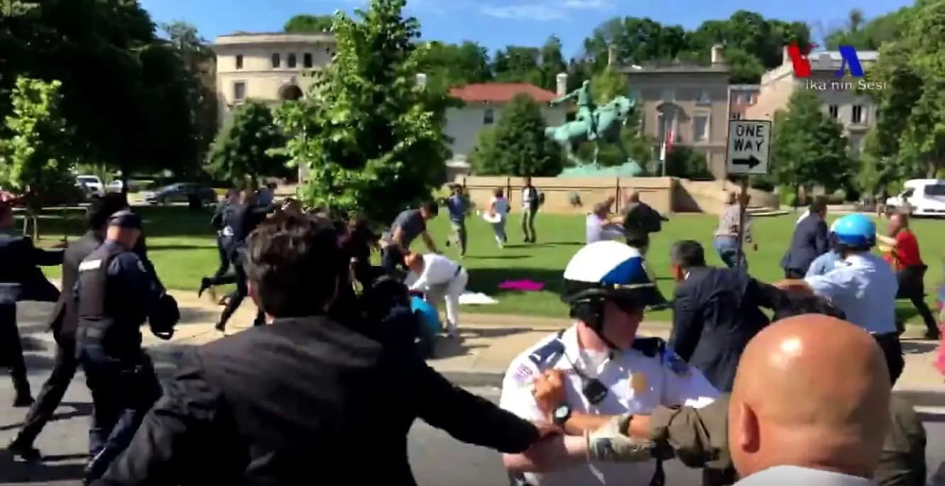 A chaotic scene on a city street shows police officers and civilians in confrontation near a park. Some people are on the ground, others are running, and officers are intervening amid trees and nearby buildings.