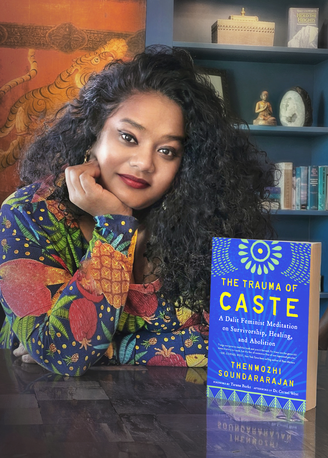 A woman with long, curly dark hair wearing a colorful patterned dress sits at a table, resting her head on her hand. In front of her is the book The Trauma of Caste by Thenmozhi Soundararajan. Shelves and art are in the background.