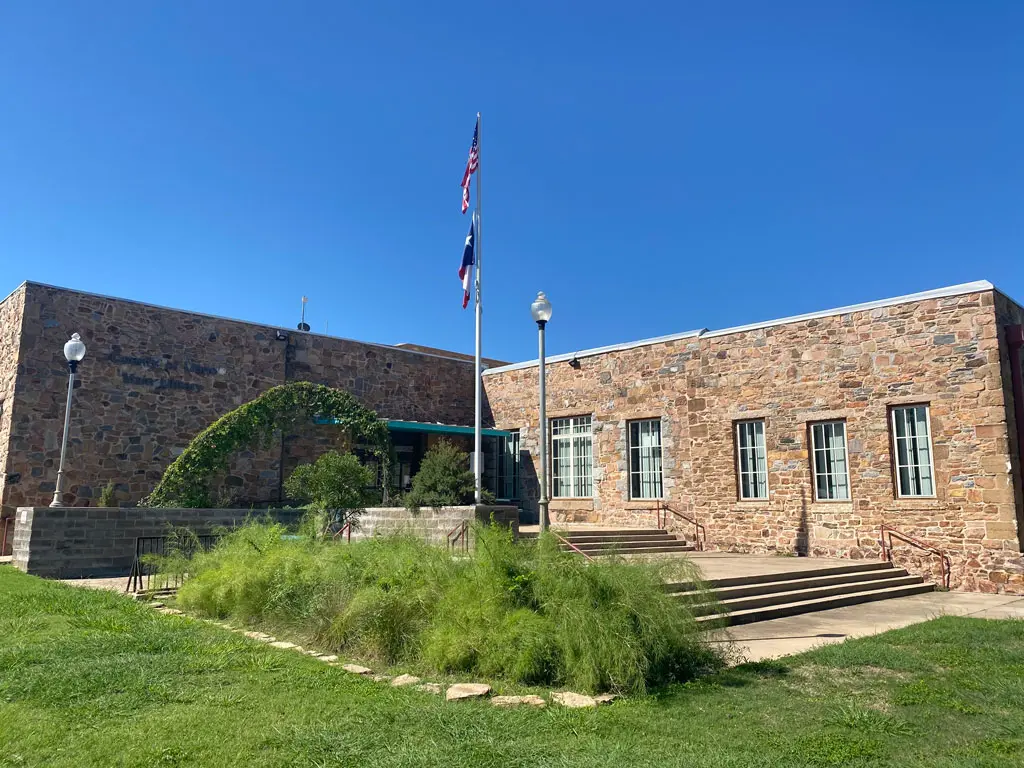 A stone building with large windows and stairs at the entrance, three flagpoles flying flags, green grass, and plants in the foreground under a clear blue sky.