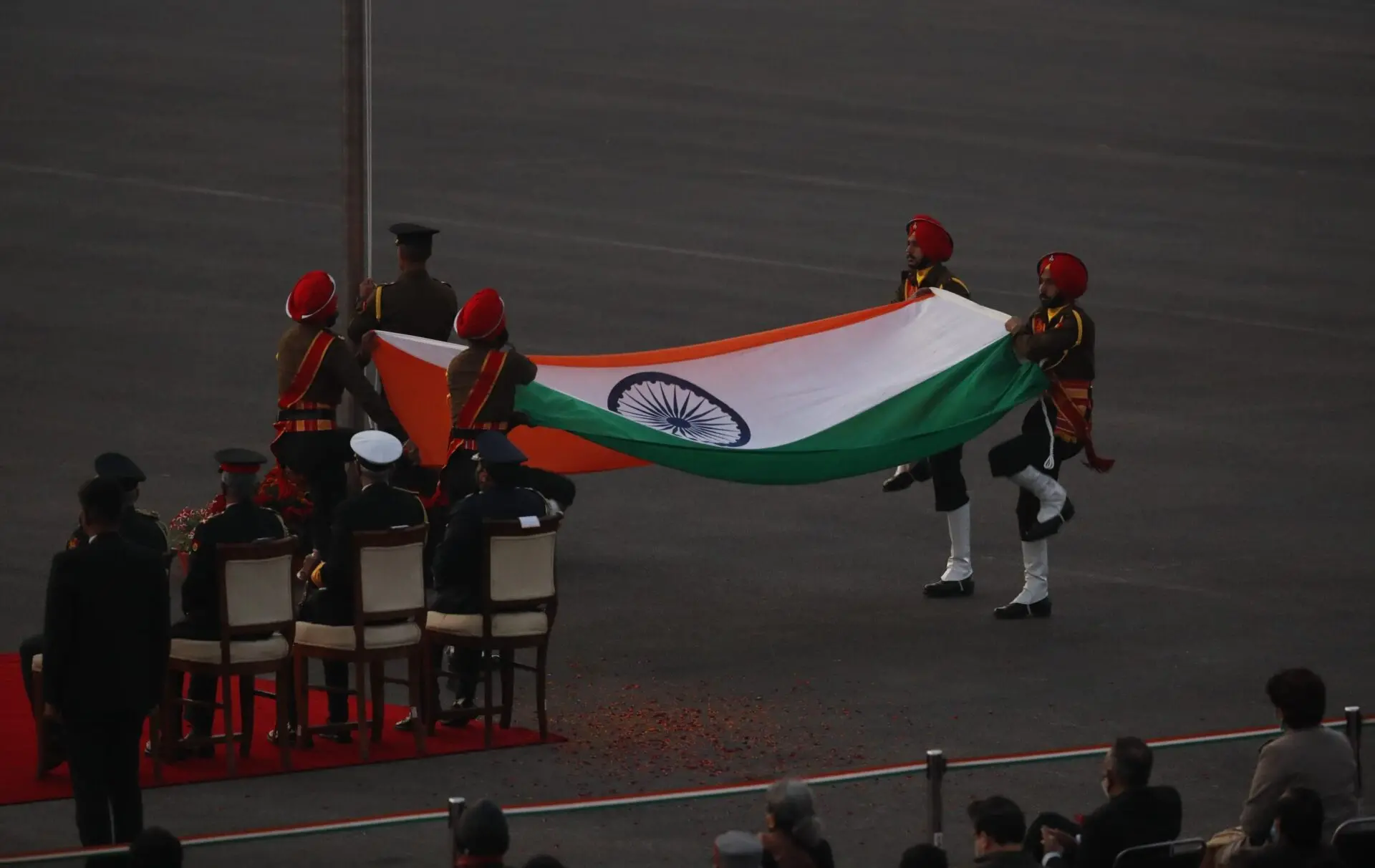 Six uniformed soldiers carry a large Indian flag during a formal ceremony, with seated officials and onlookers in the background. The scene takes place on a paved open ground.