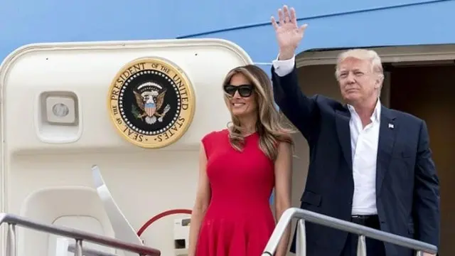 A man in a suit waves while standing next to a woman in a red dress at the entrance of an airplane displaying the presidential seal of the United States.