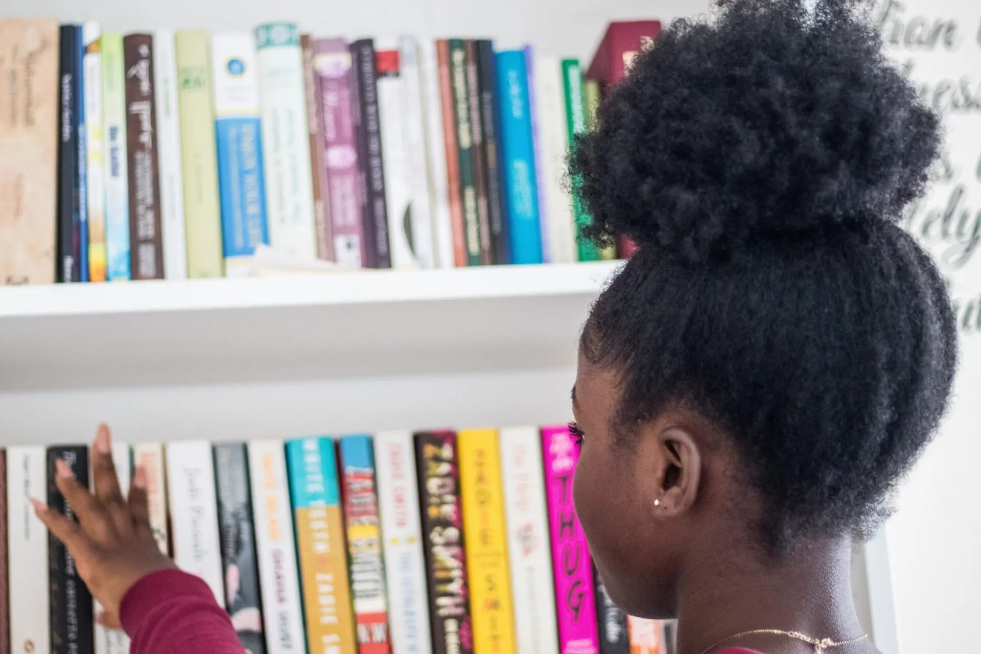 A person with natural curly hair in a puff reaches for a book on a white bookshelf filled with colorful books, viewed from behind and slightly to the side.
