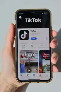 A hand holds a smartphone displaying the TikTok app page on the App Store, showing the TikTok logo, app rating, and preview images of the app interface.