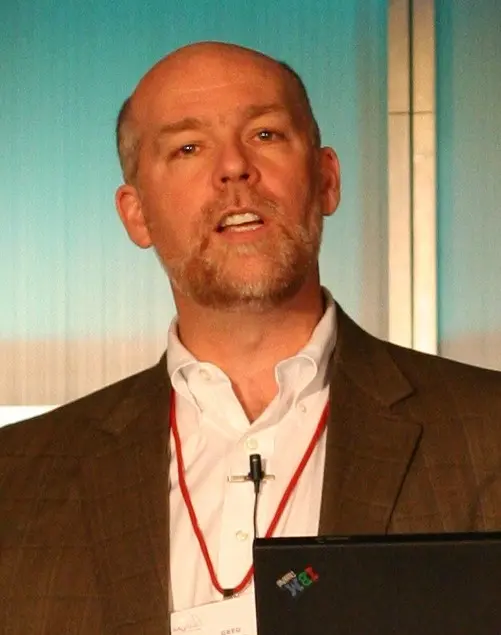A man with a trimmed beard and bald head, wearing a brown blazer, white shirt, and red lanyard, speaks in front of a blue background with a microphone clipped to his lapel.