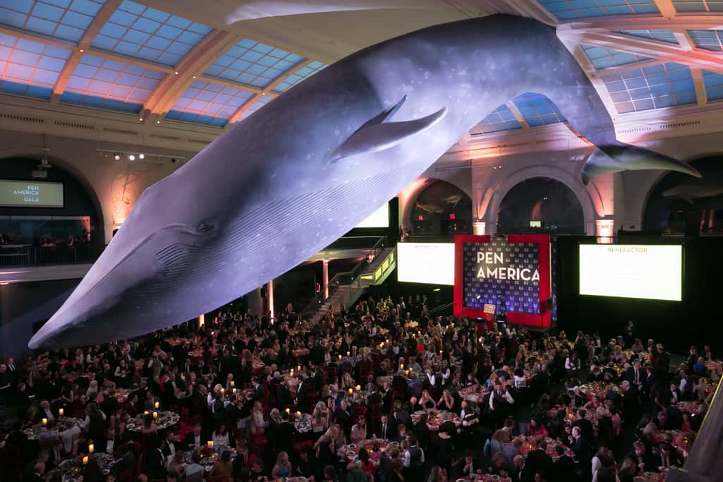A large crowd attends a PEN America event in a grand hall with a massive blue whale model suspended from the ceiling above the audience, highlighting the vibrant community passionate about PEN Americas mission.