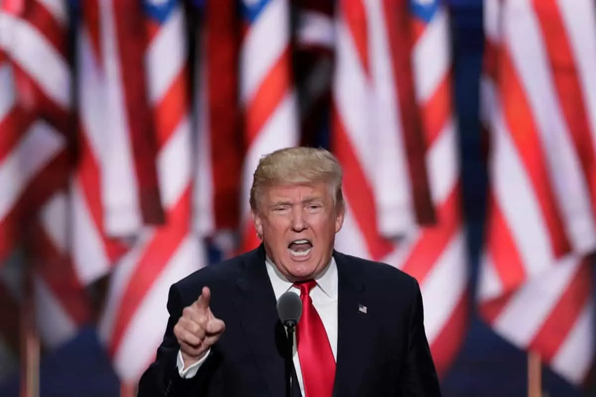A man in a suit and red tie speaks passionately at a podium, gesturing with one hand, as he addresses concerns about the coronavirus. Multiple American flags are displayed in the background.