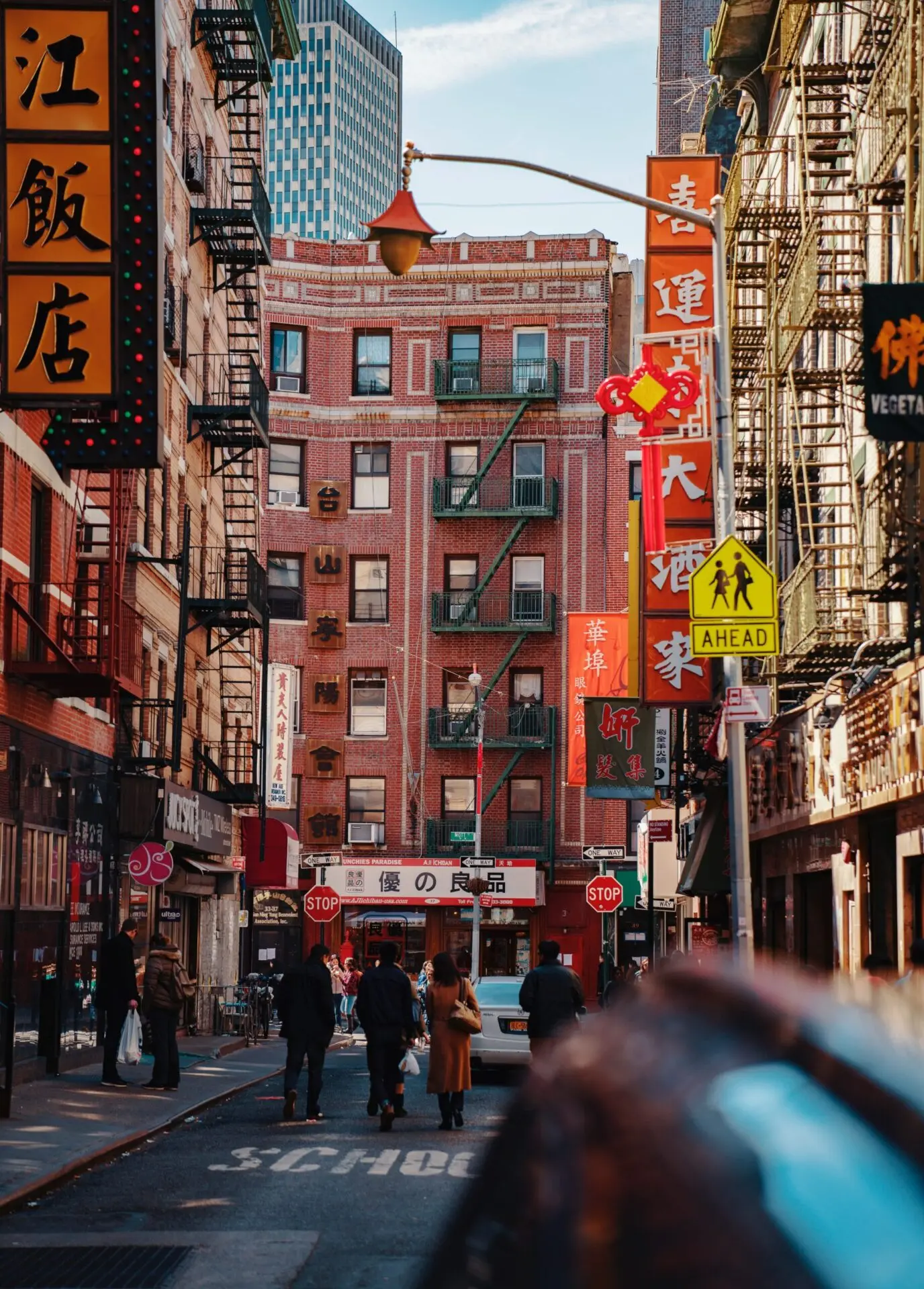 A city street scene in Chinatown with people walking, red brick buildings, fire escapes, and numerous colorful signs with Chinese characters, under a blue sky with modern skyscrapers in the background.