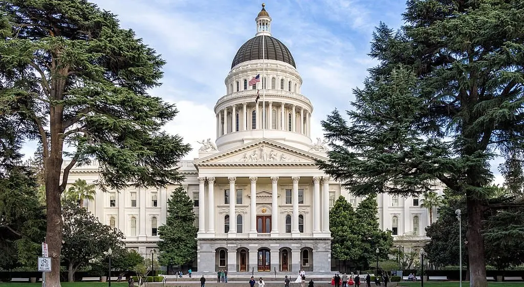 The California State Capitol building in Sacramento, featuring a large white dome, columns, and American flag, framed by tall trees, with people walking in front.