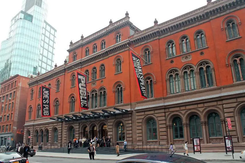 A large red-brick building with arched windows and banners reading JOES PUB and PUBLIC hanging outside. People are gathered at the entrance along a city street with modern glass buildings in the background.