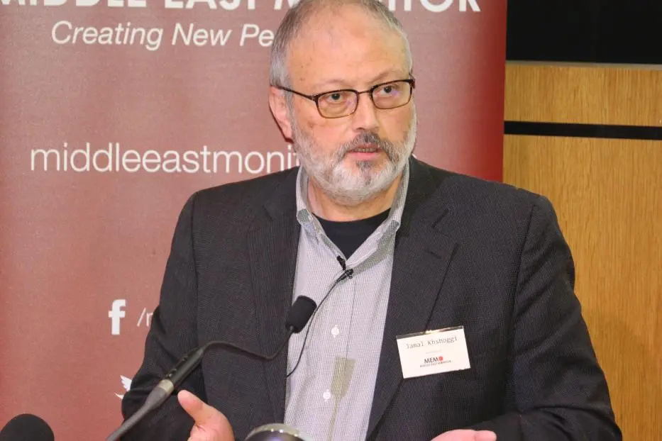 A man with glasses and a gray beard, wearing a dark blazer and striped shirt, speaks at a podium in front of a Middle East Monitor banner.