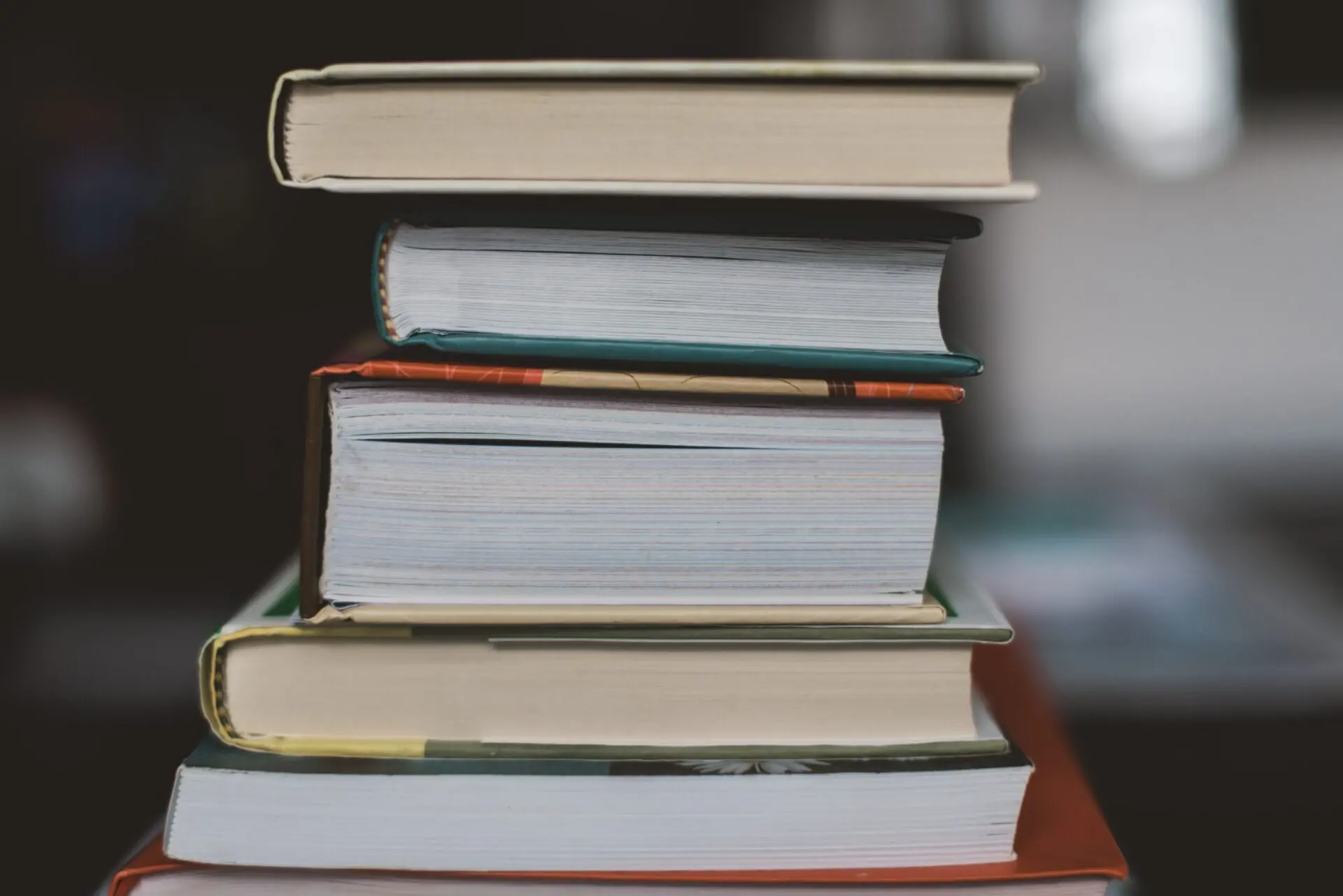 A close-up side view of a stack of five hardcover books arranged unevenly on a table, with blurred background.