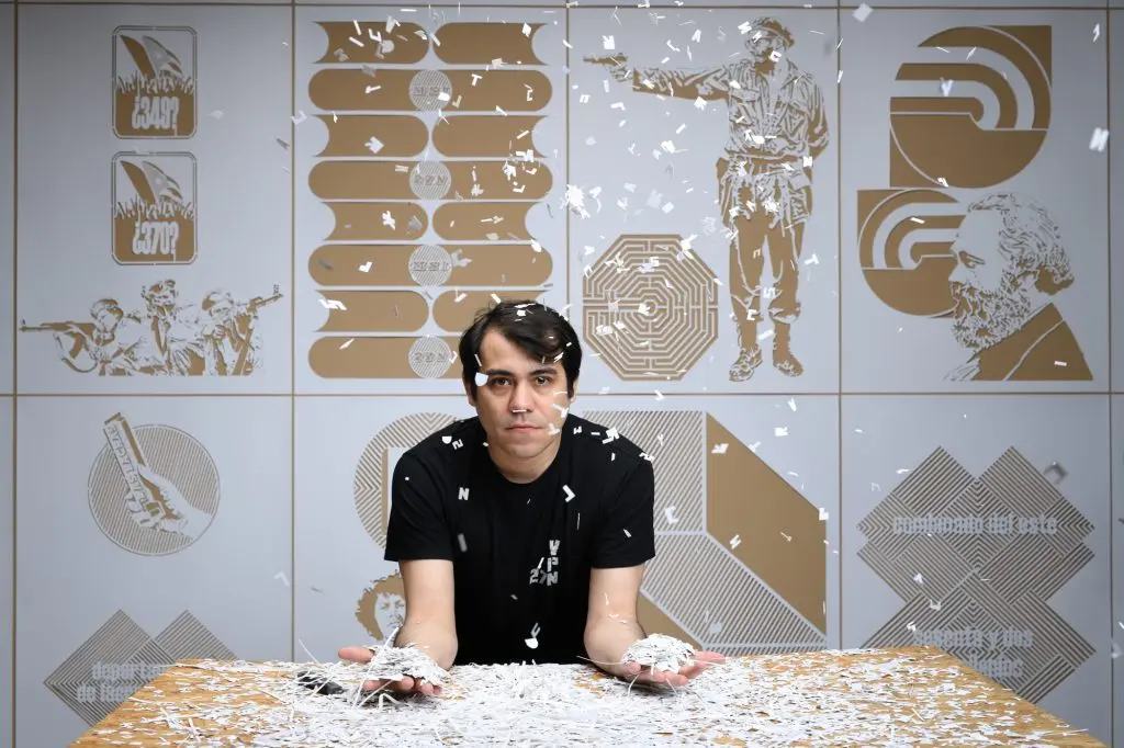 A man sits at a table holding shredded paper, with more paper falling around him. The background features stylized gold and white illustrations of people, symbols, and abstract designs.
