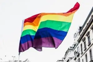 A rainbow-colored LGBTQ+ pride flag waves against a bright sky, with parts of buildings visible below on either side.