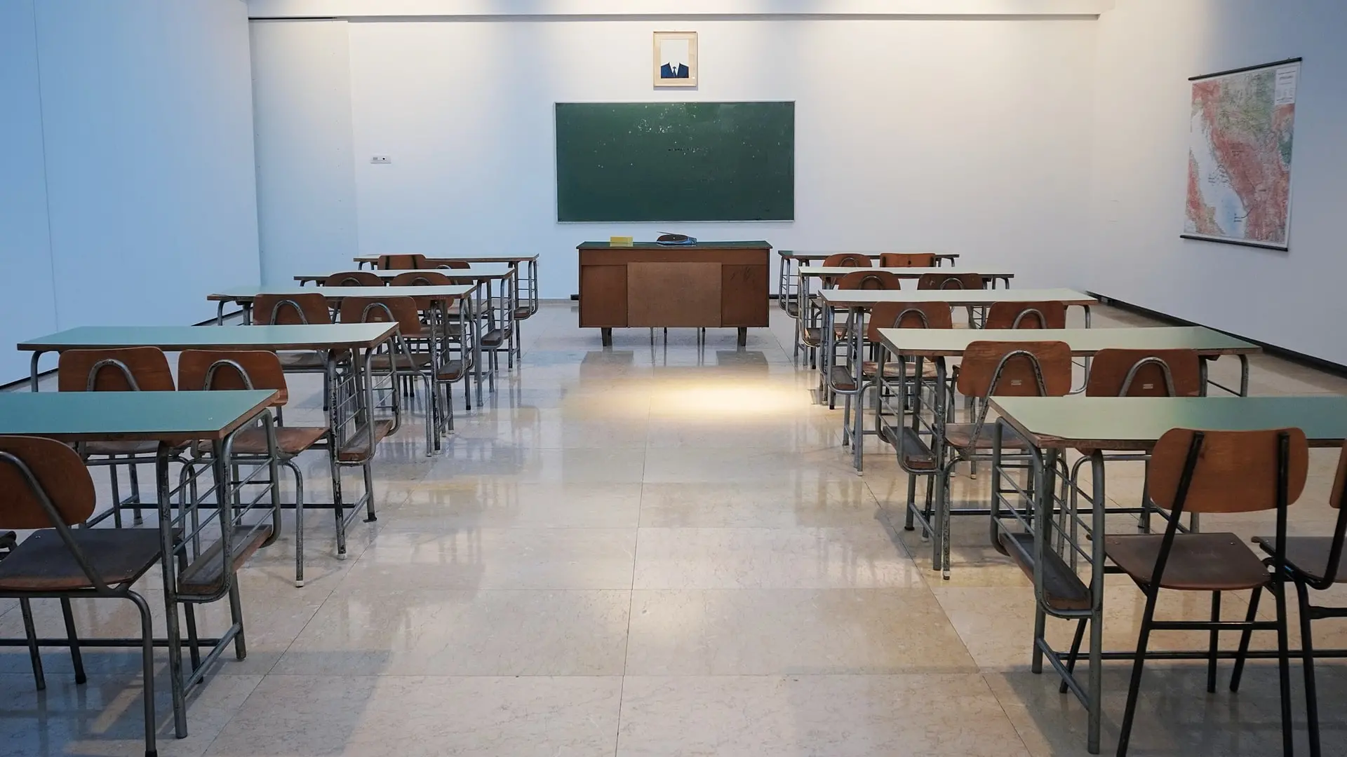 An empty classroom with rows of desks and chairs facing a teacher’s desk and a chalkboard evokes the quiet left behind when education is paused—like during coronavirus closures. A map and a framed picture hang on the tiled wall.