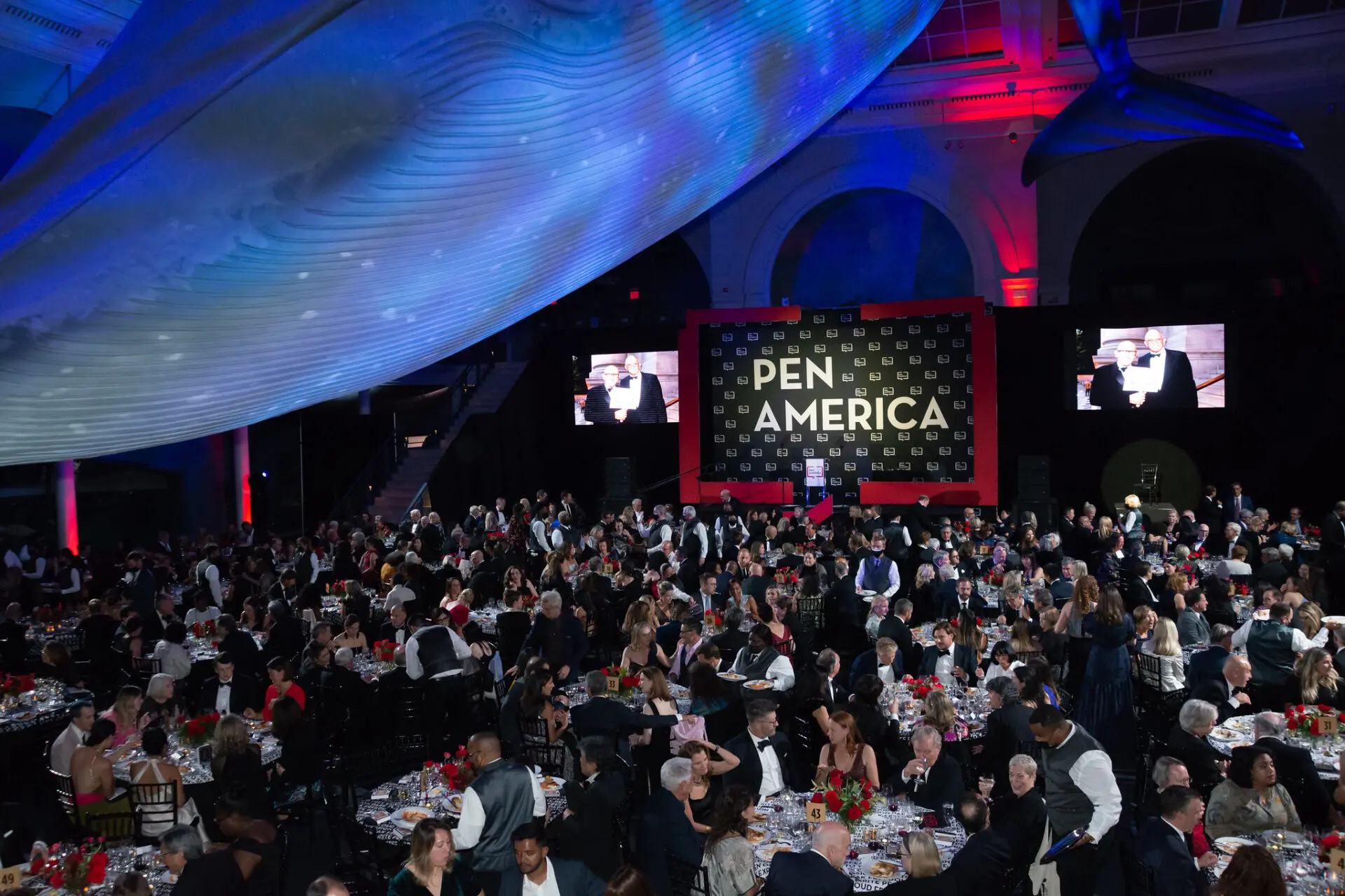 A large crowd attends a formal PEN America event in a grand hall, with tables set for dinner under a dramatic blue whale model and red-blue lighting. A stage with screens and the PEN America logo is visible in the background.