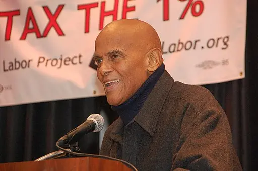 A man smiles while speaking at a podium. Behind him is a banner that reads TAX THE 1% and partially displays the web address Labor.org.