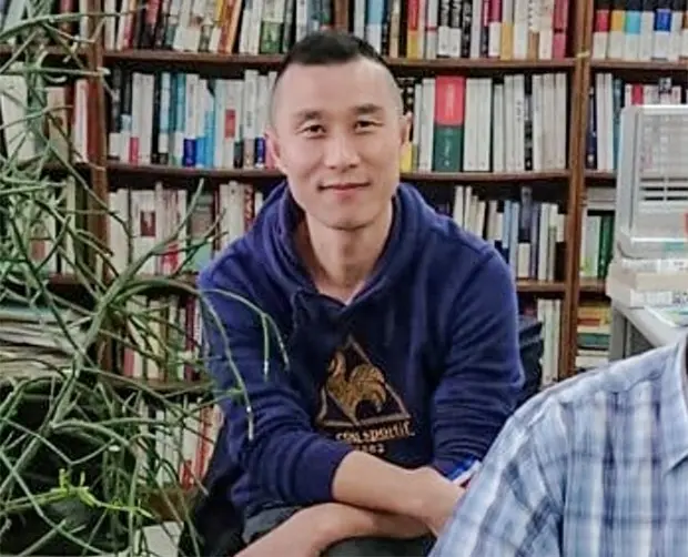 A person wearing a blue hoodie sits with arms crossed in front of bookshelves filled with books and a leafy plant; another person in a plaid shirt is partially visible in the foreground.