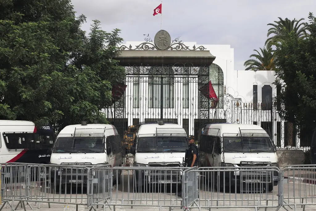 Three police vans are parked in front of a gated building with the Tunisian flag flying above. Metal barricades and officers stand in front of the entrance, with trees partially visible on either side.