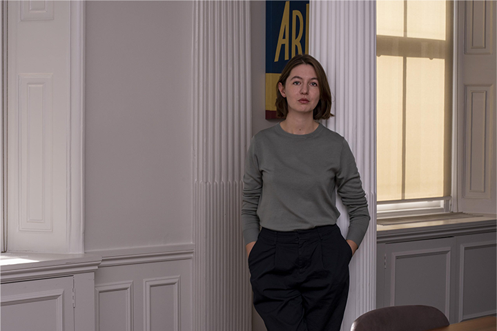 A woman with short brown hair, wearing a gray long-sleeve shirt and dark pants, stands with her hands in her pockets next to a white column in a softly lit room with large windows.