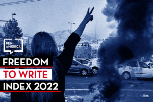 A person faces away, raising a peace sign near a cloud of dark smoke in a parking lot. Text on the image reads: “Freedom to Write Index 2022” and shows the PEN America logo. The image has a blue monochrome filter.