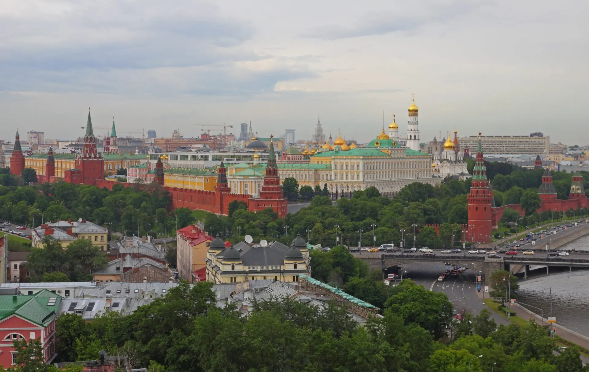 A panoramic view of the Moscow Kremlin with its red walls, golden-domed churches, and surrounding city buildings, alongside the Moskva River under a cloudy sky.