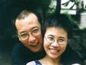A man and a woman, both wearing glasses, smile happily while posing closely together outdoors. There are blurred trees and a building in the background.