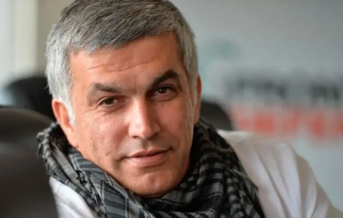 A middle-aged man with short gray hair, wearing a black and gray checkered scarf and a white shirt, looks at the camera and smiles slightly. The background is softly blurred.